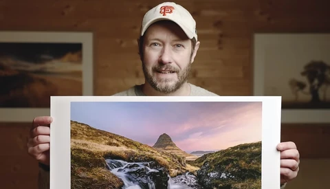Photographer displaying a landscape print of a mountain peak with stream and valley at golden hour.