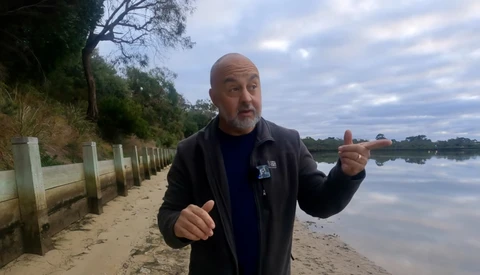 Man gesturing while speaking on a sandy beach beside calm water.