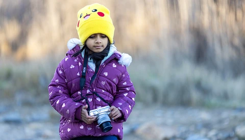 Child in purple jacket and yellow Pikachu beanie holding a camera outdoors.