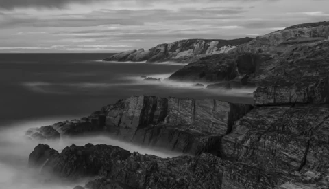 Rugged coastal cliffs meet misty ocean waters under stormy skies in black and white.