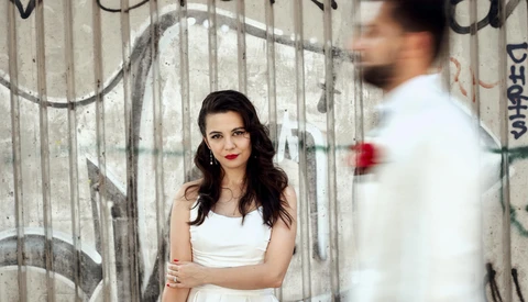 Woman in white tank top standing against graffitied concrete wall, looking at camera while blurred man in white shirt stands to her right.