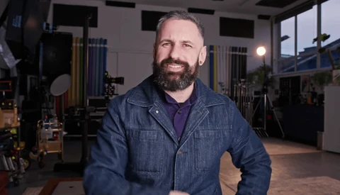 Bearded man in denim jacket smiling in a professional studio setting with lighting equipment visible.