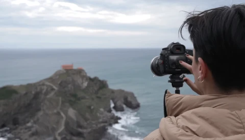 Photographer with DSLR camera on tripod capturing coastal landscape with rocky outcrop and lighthouse.