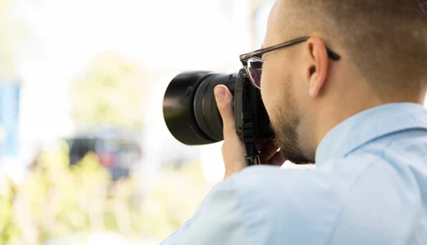 Photographer wearing glasses looks through camera viewfinder while shooting outdoors.