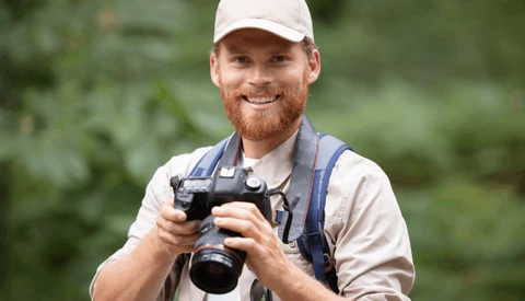 Smiling photographer in white cap holding DSLR camera outdoors with blurred green foliage background.