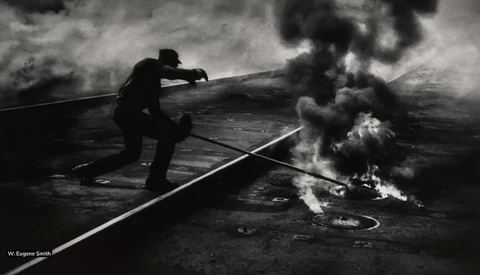 Silhouetted firefighter directing water stream at industrial fire in black and white photograph.
