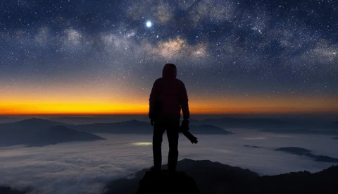 Silhouetted photographer standing on a mountain peak at twilight, facing a starry sky with the Milky Way visible above an orange and gold sunset horizon.