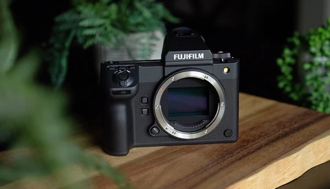 Fujifilm mirrorless camera body without lens, displayed on wooden surface with green foliage in background.
