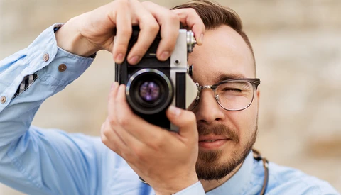 Photographer wearing glasses holding vintage film camera to his eye while looking through viewfinder.