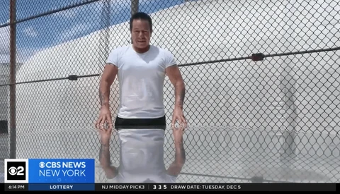 Man in white t-shirt and light pants standing against chain-link fence with blue sky background.