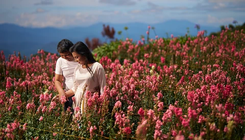 Couple in white clothing standing together in a field of blooming red flowers with mountains visible in the background.