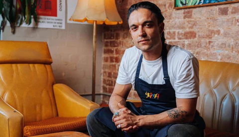 Man in dark apron seated on mustard-yellow leather chair against exposed brick wall.