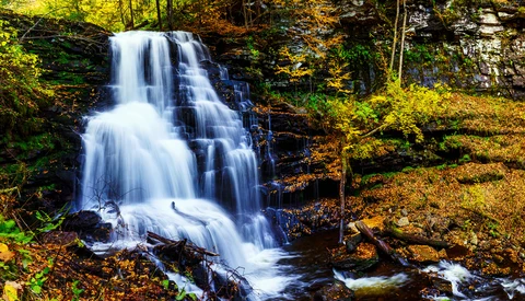 Cascading waterfall flows over rocky ledges surrounded by autumn foliage and moss-covered stone.