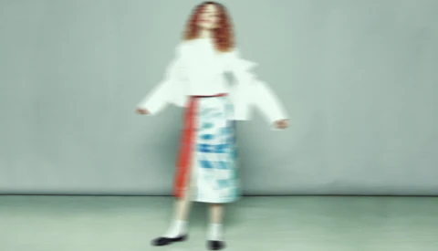 Woman in white cardigan and blue patterned skirt poses with arms extended against neutral background.