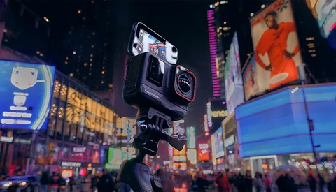 Action camera mounted on tripod in crowded urban plaza at night with illuminated billboards surrounding it.