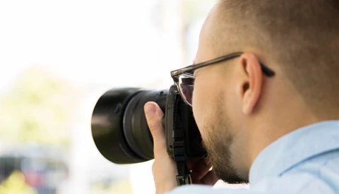 Photographer wearing glasses holding a DSLR camera to his face while shooting outdoors.