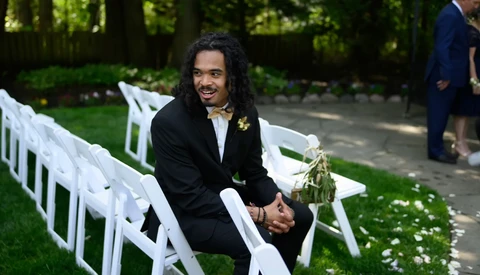Groom in black suit smiling while seated on white chair at outdoor wedding ceremony.