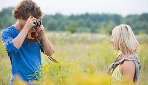 Young photographer holding vintage camera while photographing blonde woman in wildflower field.