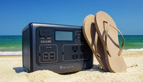 Bluetti EB55 portable power station on beach sand with flip-flops and ocean backdrop.