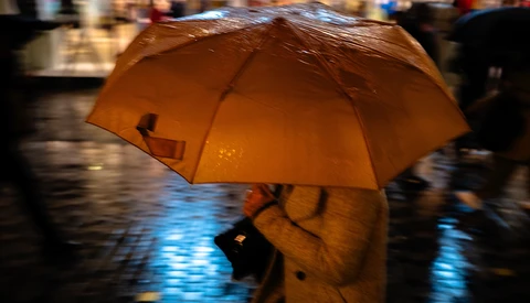Person holding a golden-orange umbrella on a wet street at dusk, with blurred pedestrians and reflective pavement in the background.