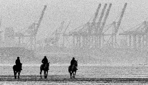 Three silhouetted figures on horseback facing an industrial waterfront with cranes and shipping infrastructure shrouded in mist.
