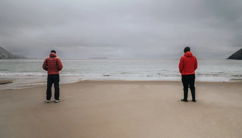 Two people in red jackets standing separately on a beach facing the ocean under overcast skies.