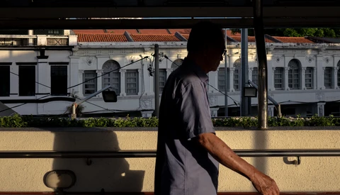 Silhouetted figure leaning against a railing with colonial-style buildings visible through glass in bright sunlight.