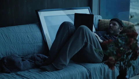 Person lounging on bed in dim blue-toned light while working on laptop and tablet.
