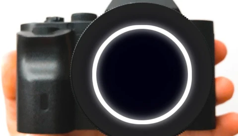Compact camera with black body and orange grip held in hand, featuring illuminated circular lens ring.