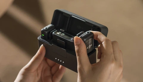Hands holding open a charging case containing two wireless microphone transmitters against a neutral beige background.