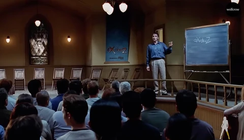 Man presenting to seated audience in wood-paneled room with blue whiteboard behind him.
