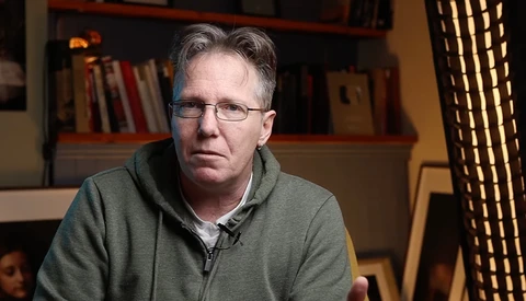 Man wearing glasses and gray hoodie sitting indoors with bookshelves in background.