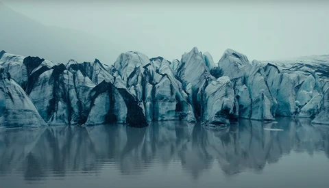 Jagged glacier face with dark crevasses reflected in still water below.