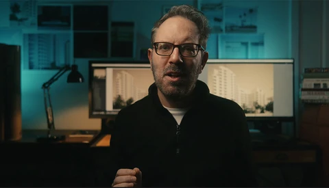 Man with glasses speaking in a studio with dual monitors and teal-blue ambient lighting.