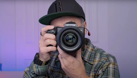 Photographer holding a Fujifilm medium format camera up to eye level, wearing a black baseball cap and plaid shirt.