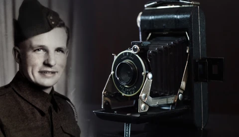 Man in military uniform posing beside a large format folding camera on a stand.