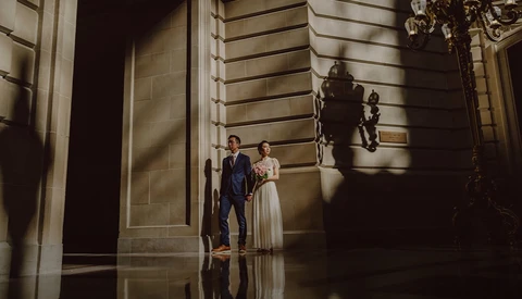 Bride and groom standing in a classical hallway with dramatic side lighting casting shadows on stone walls.