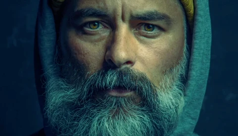 Bearded man with intense gaze wearing a teal hood against dark background.
