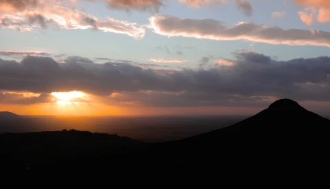 Silhouetted volcanic cone against a golden sunset with dramatic cloud formations and warm orange light breaking through the sky.