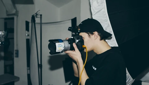 Photographer in black cap and shirt holding a professional video camera with yellow shoulder strap in a studio setting.