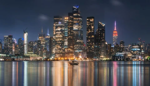 Illuminated Manhattan skyline at dusk reflected in calm river water.