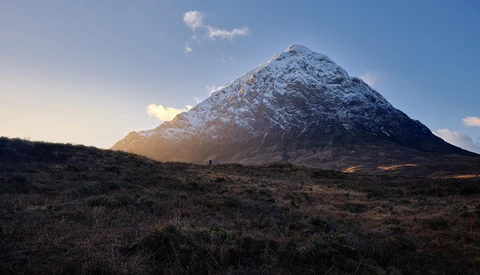 Snow-capped conical mountain rising from barren moorland under clear blue sky.