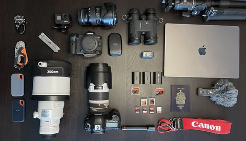Flat lay of professional photography gear including Canon DSLR camera, lenses, MacBook, binoculars, memory cards, and accessories arranged on dark surface.