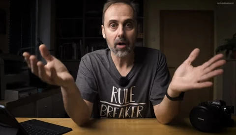 Man in a dark v-neck shirt gesturing expressively while speaking to camera indoors.