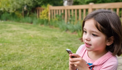 Young girl in pink shirt looking down at smartphone in backyard setting.