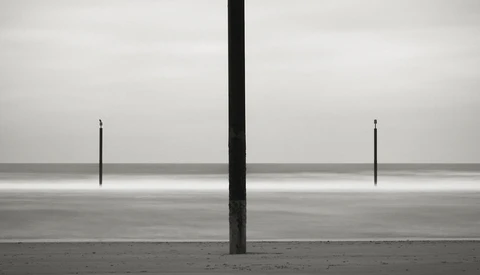 Three vertical posts on a vast, flat beach with calm water and overcast sky in stark monochromatic composition.