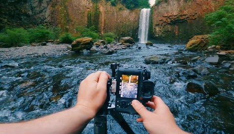 Photographer's hands holding a camera displaying a waterfall scene in the LCD screen, with the actual waterfall and rocky stream visible in the background.
