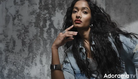 Woman with long dark hair posing against a textured gray wall, hand raised to face.