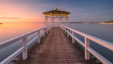 Wooden pier with white railings extending toward a gazebo at sunrise over calm water.
