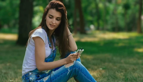 Young woman in denim overalls and white t-shirt sitting outdoors holding a smartphone.